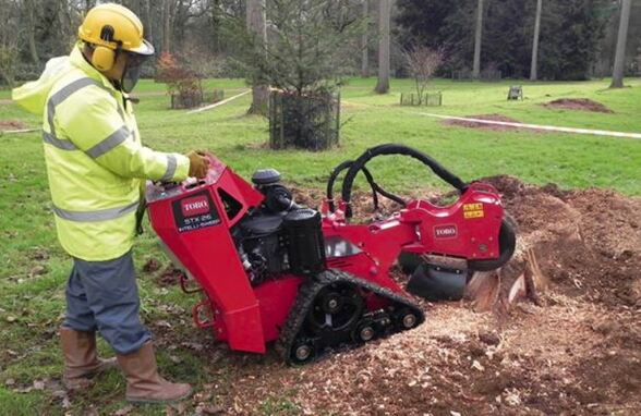  A man is standing behind a stump grinding machine and removing a stump. He's dressed in safety gear from head to toe. Has ear and eye protection as well. The background is green grass and a Forrest. 